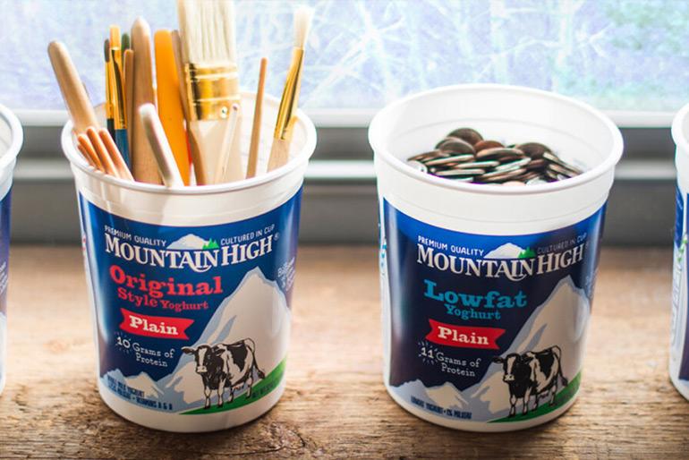 Two pots of Mountain High Yoghurt that have been repurposed, one into a paintbrush holder and the other into a makeshift piggy bank filled with coins.