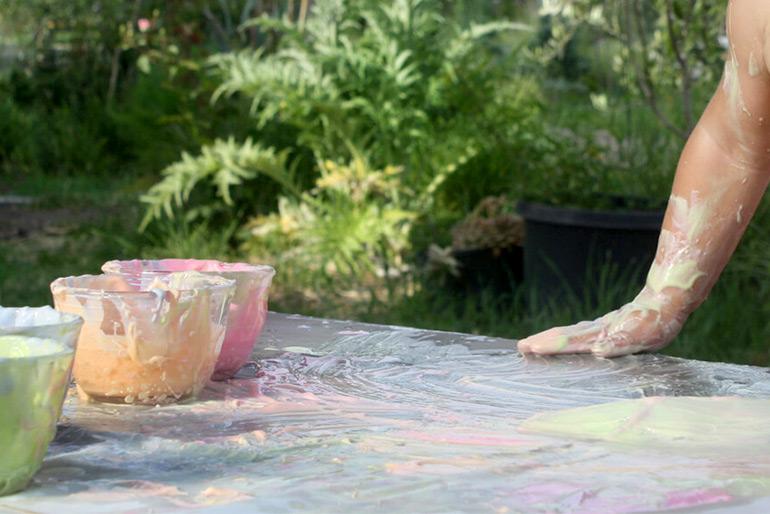 A person's hand taking part in finger painting with the Yoghurt Finger Paints in bowls to one side and a large surface being used as the canvas.